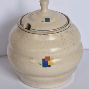 May include: A round, off-white ceramic sugar bowl with a speckled finish and a matching lid. The bowl features a geometric pattern of small blue, red, and yellow squares with thin green lines. It has a ribbed body and a small notch for a spoon.