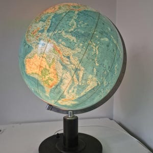 以下が含まれることがあります： ターコイズとタン色の地図デザインが施された、ヴィンテージの照明付き地球儀ランプ。地球儀は、円筒形の支柱と丸いベースを備えた黒いベースに取り付けられています。ランプはプラグインされており、点灯して大陸を表示しています。