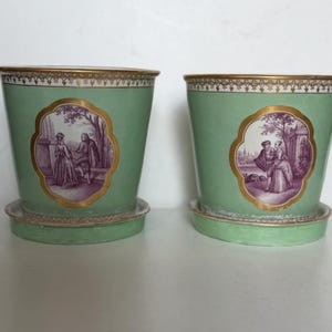 Peut inclure: Deux cache-pots en céramique vert clair avec soucoupes assorties, dotés d'un bord doré et d'un cartouche central contenant une illustration monochrome violette de personnages dans un paysage, sur un fond blanc uni.