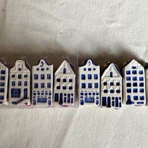 Op de afbeelding: Een set van acht miniatuur huisjes van keramiek, elk met een witte basis en handbeschilderde blauwe architectonische details zoals ramen en deuren. Vier van de huisjes zijn gerangschikt in een rechthoekige, doorzichtige plastic verpakking.