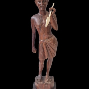 Könnte beinhalten: Eine geschnitzte Holzstatue einer stehenden Figur, die einen Speer hält. Die Figur ist braun mit einem einfachen Lendenschurz und einer Kopfbedeckung. Der Speer ist weiß. Die Statue ist auf einem Holzsockel vor schwarzem Hintergrund montiert.