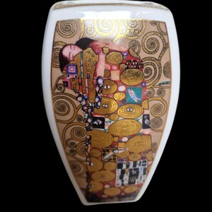 Op de afbeelding: Een witte keramische vaas met een schilderij van Gustav Klimt. Het kunstwerk toont een vrouw in een gouden en kleurrijke jurk, omringd door gouden spiraalpatronen. De vaas heeft een afgeronde vorm met een smalle hals.
