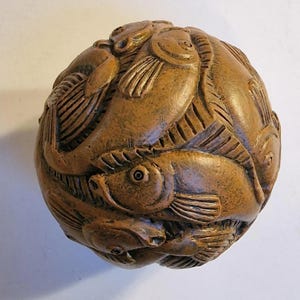 Op de afbeelding: Een houten bol met een gedetailleerd reliëf van meerdere vissen. De vissen zijn gerangschikt in een wervelend patroon, met zichtbare vinnen, schubben en ogen. Het hout heeft een warme, bruine tint en de bol is waarschijnlijk een decoratief item.