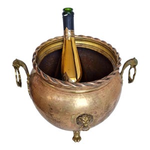 Op de afbeelding: Een gouden champagnefles in een vintage messing ijsemmer. De ijsemmer heeft een decoratieve touwachtige rand rond de rand, handvatten en een leeuwenkopdetail. De fles is gedeeltelijk zichtbaar, met een zwart en goud label.