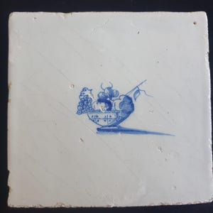 May include: Square, white ceramic tile with a blue and white illustration of a bowl overflowing with fruit. The fruit includes grapes and other stylized elements. The tile has a slightly distressed appearance.