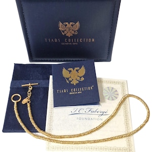 Op de afbeelding: Een goudkleurige ketting met een T-bar sluiting en een kleine ring, gepresenteerd met een donkerblauwe leren etui en een blauw fluwelen zakje. De etui en het zakje hebben gouden letters en een gouden embleem. Een certificaat is ook inbegrepen.