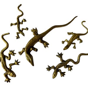 Puede incluir: Una colección de cinco figuras de lagartos de latón en varios tamaños y poses. Cada lagarto presenta una superficie metálica de tono dorado texturizada con escamas detalladas, cuatro patas extendidas con dedos visibles y una cola larga y curva.