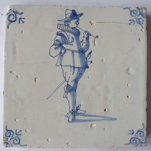 Könnte beinhalten: Quadratische Keramikfliese mit einer blau-weißen Illustration eines Mannes in historischer Kleidung. Die Figur trägt einen Hut und hält einen langen Gegenstand an den Mund. Blaue Wirbel zieren die Ecken. Das Design wirkt antik.