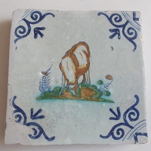 May include: Square ceramic tile with a hand-painted scene of two brown and white animals grazing on green grass. The tile has a light blue background with blue scrollwork in the corners.