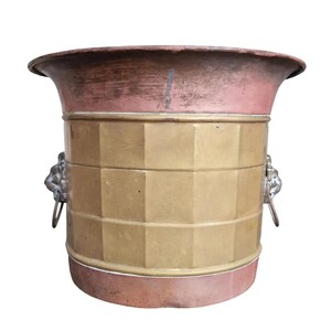 Op de afbeelding: Een koperen en messing emmer met een uitlopende rand en een geometrisch patroon. De emmer heeft een messing middengedeelte met een geometrisch ontwerp en koperen boven- en onderdelen. Leeuwenkop handvatten zitten aan weerszijden.