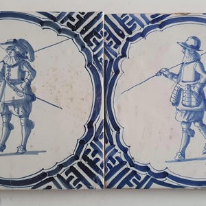 May include: Two antique ceramic tiles with blue and white illustrations of soldiers. Each figure holds a long pole and wears period clothing. The tiles have a decorative blue border.
