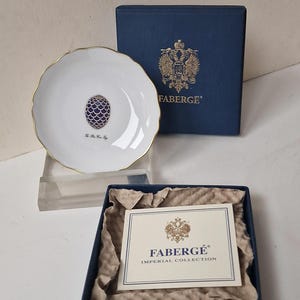 Op de afbeelding: Een witte Fabergé-bord met een gouden rand en een centraal blauw eierontwerp. De plaat wordt getoond met de marineblauwe doos, met het Fabergé-logo en tekst. Een kaart uit de Imperial Collection is ook zichtbaar.