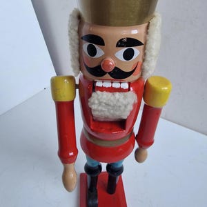 Op de afbeelding: Een houten notenkrakerfiguur met een goudkleurige hoed, een rode jas en een zwarte snor. Het beeldje heeft een witte baard en staat op een rode basis. De armen zijn rood met gele manchetten.