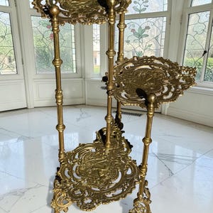 May include: A tiered gold-toned metal plant stand featuring three circular shelves with intricate openwork filigree patterns and embossed central motifs. The structure is supported by slender, segmented vertical columns and ornate, curved legs.