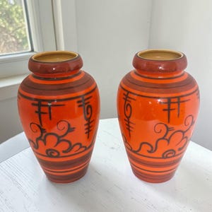 Puede incluir: Dos jarrones de cerámica naranja con diseños decorativos marrones y negros. Los jarrones tienen forma redondeada con cuello estrecho y base ancha. El diseño incluye rayas horizontales, símbolos estilizados y motivos de nubes.