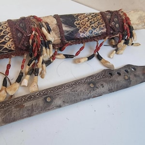 Puede incluir: Un cuchillo antiguo con una vaina decorativa. La vaina está envuelta en cuero estampado y adornada con borlas de cuentas rojas, negras y blancas. La hoja es de metal con una serie de agujeros y diseños grabados.