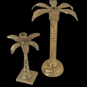 Könnte beinhalten: Zwei goldfarbene Palmen-Kerzenhalter. Einer ist hoch mit einem geriffelten Stamm und einem runden Sockel mit Rautenmuster. Der andere ist kürzer mit einem quadratischen Sockel. Beide haben detaillierte Palmwedel.