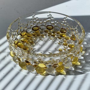 Puede incluir: Un cuenco de vidrio transparente con un patrón decorativo de formas de burbujas ámbar y transparentes. El cuenco redondo tiene un borde ligeramente ensanchado y se asienta sobre una base transparente. Las burbujas varían en tamaño y color.