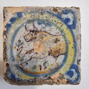 Pode incluir: Azulejo quadrado antigo com um desenho central que apresenta uma figura animal estilizada em castanho e amarelo. O animal é circundado por um anel amarelo e azul, com detalhes florais azuis nos cantos. O azulejo tem um aspeto envelhecido.