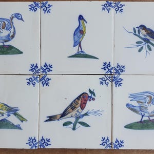Può includere: Sei piastrelle di ceramica quadrate con illustrazioni di uccelli dipinte a mano. Ogni piastrella presenta un diverso disegno di uccello in blu, giallo e marrone, su sfondo bianco. Gli angoli di ogni piastrella hanno un motivo floreale blu.