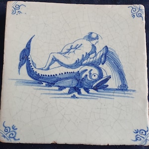 Może przedstawiać: Kwadratowa płytka ceramiczna z białym tłem i niebieską, ręcznie malowaną ilustracją postaci jadącej na dużej rybie. Wzór posiada wykończenie z popękaną glazurą oraz dekoracyjne niebieskie motywy w rogach płytki.