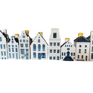 May include: A collection of eight ceramic miniature house figurines arranged in a row. Each piece features a white base with blue painted architectural details, including windows, doors, and gabled roofs, topped with small yellow cylindrical stoppers.