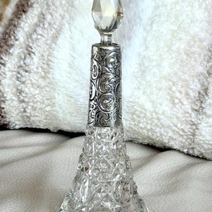 May include: A clear glass perfume bottle with a conical base featuring a diamond-cut pattern. The neck is adorned with a silver-colored metal collar displaying an intricate embossed floral scroll design, topped by a faceted glass stopper.