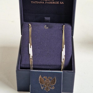 Puede incluir: Pendientes dorados con detalles blancos, presentados en una caja azul marino. La caja está abierta, mostrando los pendientes contra un interior azul oscuro. Una pequeña tarjeta con el texto "TSARS COLLECTION" está al frente.