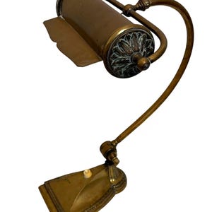 Könnte beinhalten: Eine Schreibtischlampe aus Messing mit einem zylindrischen Schirm, einer dekorativen floralen Endkappe, einem gebogenen verstellbaren Arm und einem rechteckigen Sockel. Die Lampe verfügt über einen kleinen weißen Druckschalter auf der Oberseite des Sockels und weist eine verwitterte Metalloberfläche auf.