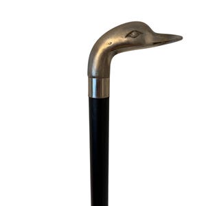 Op de afbeelding: Een wandelstok met een zilverkleurig metalen handvat in de vorm van een eendenkop. Het handvat is bevestigd aan een gladde, rechte, zwarte houten schacht met een gepolijste zilverkleurige metalen kraag die de kop met de donkere stok verbindt.