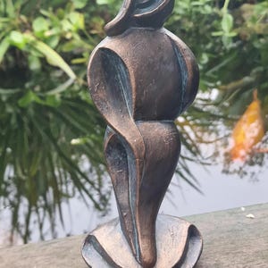 May include: Bronze-colored abstract sculpture of a person in a flowing pose. The figure has a smooth, curved design and is mounted on a rounded base. The sculpture is set against a blurred background of greenery and water.