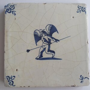 Op de afbeelding: Vierkante keramische tegel met een crèmekleurige achtergrond en een blauwe illustratie van Cupido die een pijl richt. De tegel heeft een gebarsten uiterlijk en decoratieve blauwe hoekaccenten.