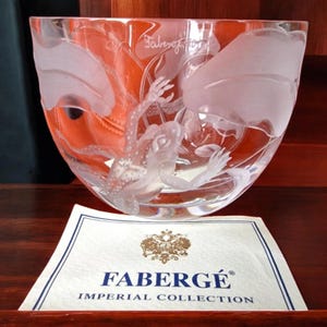 Op de afbeelding: Een heldere glazen kom met geëtste kikker- en waterlelie-ontwerpen. De kom heeft een brede opening en een gebogen vorm. De basis van de kom heeft een wit label met de tekst "FABERGÉ IMPERIAL COLLECTION".