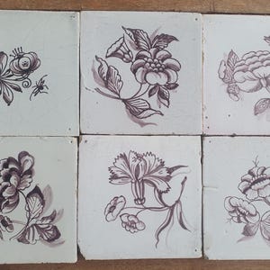 Op de afbeelding: Een set van zes vierkante keramische tegels met een witte basis en donkerpaarse bloemillustraties. Elke tegel heeft een uniek botanisch ontwerp, inclusief diverse bloemen en kleine insecten, met zichtbare haarscheurtjes in het glazuur.