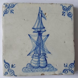 Op de afbeelding: Vierkante keramische tegel met een handgeschilderd blauw zeilschipontwerp. Het schip heeft meerdere masten en zeilen, met twee grote, afgeronde vormen aan de basis. Decoratieve blauwe bloemenhoekaccenten. De tegel is gebroken wit.