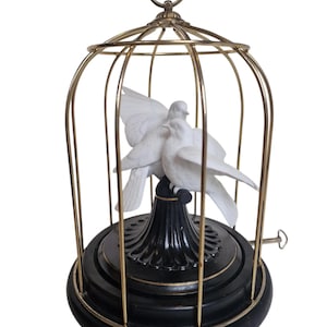 Op de afbeelding: Een decoratieve vogelkooi met een goudkleurig frame en een zwarte basis. Binnenin zitten twee witte duifjes op een zwarte standaard. De kooi heeft een handvat aan de bovenkant en een opwindsleutel aan de zijkant.