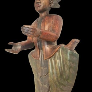 Op de afbeelding: Een gesneden houten beeld van een persoon in traditionele kledij. De figuur heeft een serene uitdrukking en uitgestrekte handen. Het beeld is voornamelijk bruin met groene en rode accenten, tegen een zwarte achtergrond.