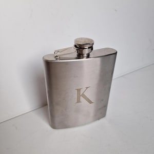 Könnte beinhalten: Eine Edelstahl-Flachmann mit Klappverschluss und Schraubverschluss. Der Flachmann hat eine gebogene Form und trägt den Buchstaben "K" auf der Vorderseite. Der Flachmann ist zur Aufnahme von Flüssigkeiten bestimmt.