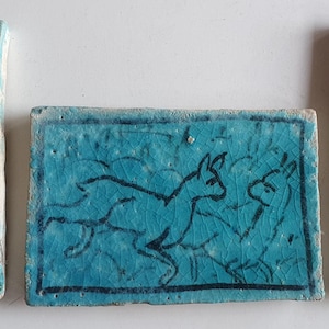 Könnte beinhalten: Drei rechteckige, türkisfarbene Keramikfliesen. Zwei Fliesen zeigen florale Designs in schwarzer Tinte. Die mittlere Fliese zeigt zwei Tiere in schwarzer Tinte. Die Fliesen haben ein rissiges, gealtertes Aussehen.