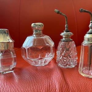 May include: Four vintage glass bottles with silver-toned metal caps and hardware. The collection includes a square bottle with horizontal lines, a round faceted bottle, a small diamond-patterned bottle with a nozzle, and a tall fluted bottle.