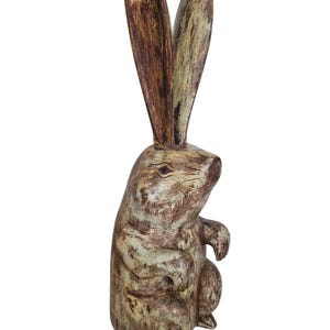 Può includere: Una statuetta di coniglio rustico in legno, con lunghe orecchie dritte. La scultura ha una finitura anticata nei toni del marrone e del crema, che le conferisce un aspetto invecchiato. Il coniglio è raffigurato in posizione seduta, con le zampe appoggiate sul petto.
