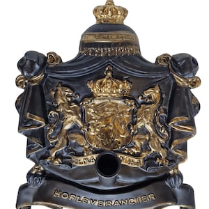 Puede incluir: Un emblema decorativo negro y dorado con un escudo de armas real con dos leones, una corona y el texto "HOFLEVERANCIER". El emblema tiene un fondo oscuro con detalles dorados y una abertura cilíndrica.