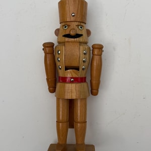 Könnte beinhalten: Eine hölzerne Nussknacker-Figur mit natürlicher hellbrauner Oberfläche, die einen hohen zylindrischen Hut trägt. Die Figur hat einen schwarz bemalten Schnurrbart, einen leuchtend roten Gürtel und silberfarbene Metallnieten an Brust und Hut.