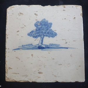 Op de afbeelding: Vierkante, gebroken witte keramische tegel met een blauw boomontwerp. De boom heeft een gedetailleerde, getextureerde kroon en een slanke stam. Het oppervlak van de tegel heeft een verweerde uitstraling, met zichtbare imperfecties en een licht ruwe textuur.