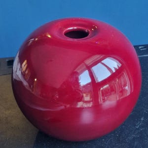 Op de afbeelding: Een glanzende, bolvormige, rode keramische vaas met een kleine opening aan de bovenkant. De vaas reflecteert de omgeving. De vaas is een decoratief item.