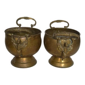 Op de afbeelding: Een paar bijpassende koperen kommen, elk met een ronde vorm, een voetstuk en twee handvatten. De handvatten zijn versierd met een reliëf van een gevleugelde figuur. Het koper heeft een verweerde, verouderde patina met zichtbare textuur.