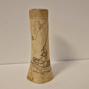 Op de afbeelding: Een beige, cilindrisch object met een gedetailleerde gravure van een visser in een boot. De visser wordt afgebeeld met een hengel en de boot bevindt zich op het water. De bovenkant van het object heeft een decoratieve, getextureerde band.