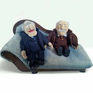 Op de afbeelding: Twee pluche poppen van oudere mannen, een in een marineblauw krijtstreep pak en de andere in een bruin pak, zitten op een lichtblauwe chaise longue. De poppen hebben wit haar, snorren en rode lippen. De chaise longue heeft een donkerhouten frame.