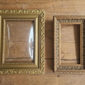 Op de afbeelding: Twee sierlijke, goudkleurige fotolijsten met ingewikkelde details. Eén lijst is rechthoekig en groter, met een heldere glazen inzet. De andere lijst is kleiner en rechthoekig, met een gedetailleerde rand. Beide lijsten zijn leeg.