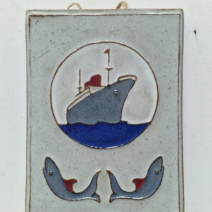 Op de afbeelding: Een rechthoekige keramische tegel met een nautisch thema. De tegel toont een blauw en grijs schip in een witte cirkel boven blauwe golven. Twee gestileerde blauwe dolfijnen met rode accenten staan eronder. De tegel heeft een lichtblauwe achtergrond en een bruine rand.